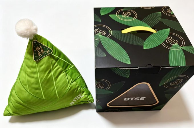 The image features a green pillow with white pom-poms, placed next to a black box that has a green leaf pattern. The pillow appears to be made of fabric or material similar to a Christmas stocking stuffer. Both objects are sitting on a table against a white background.