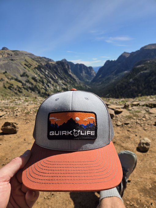 A hand holds a gray and orange trucker hat featuring a 'QUIRK LIFE' mountain patch, with a scenic mountainous landscape in the background.