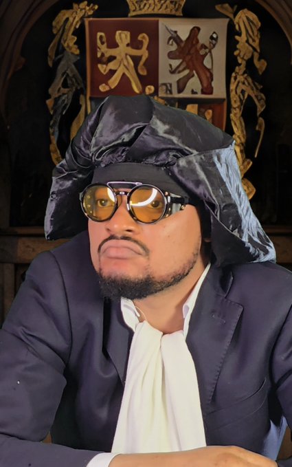 A man wearing yellow-tinted round sunglasses, a dark suit jacket over a white cravat, and a traditional black horsehair legal wig, posed in front of a heraldic crest.
