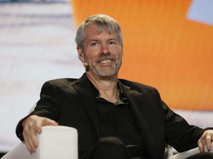 A man with gray hair and a beard, wearing a black blazer and headset microphone, speaks while seated on stage at what appears to be a conference.