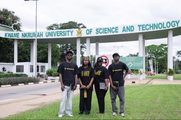 Four young people wearing matching "I BELIEVE" shirts pose together in front of the prominent entrance archway of Kwame Nkrumah University of Science and Technology (KNUST), capturing the team's visit to the campus. The image visually reinforces the post's message by showing the group actively present on-site, embodying the "community" and connection to the university mentioned in the caption.