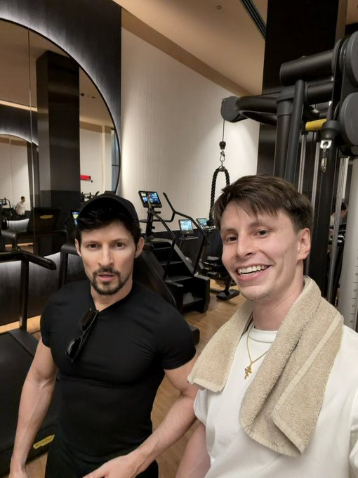 Two men taking a selfie in a modern gym setting; one wears a black cap and fitted shirt, while the other smiles wearing a white t-shirt with a towel draped over his shoulder, with exercise equipment visible behind them.