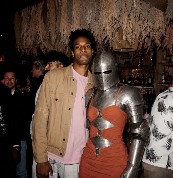 A photograph of a man in a pink shirt and tan jacket standing next to someone wearing medieval knight armor over an orange dress at what appears to be a bar or party venue.