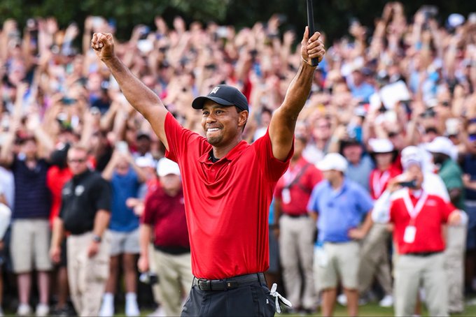 The image shows Tiger Woods in a red Under Armour shirt and black cap, raising his arms triumphantly in front of a cheering crowd, likely celebrating a major golf victory. This image is unrelated to the post about DebtReliefBot (DRB), which provides a Base network smart contract address for the cryptocurrency token.