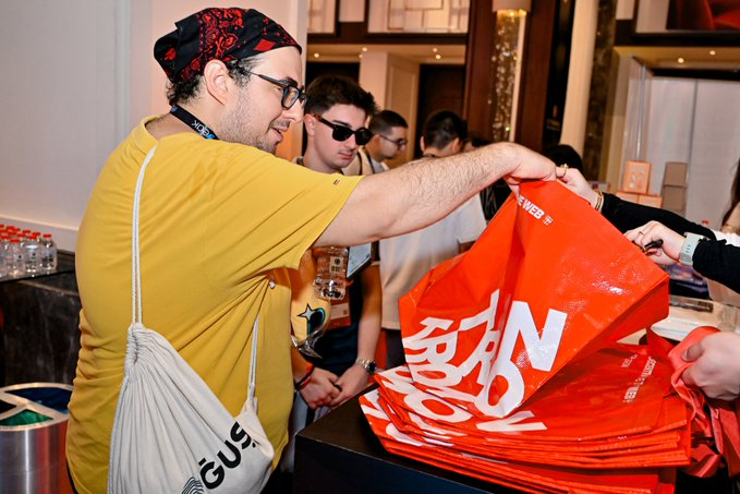 A man in a yellow shirt and red bandana distributes orange "SunSwap" tote bags at what appears to be a SunSwap V3 launch event, with another attendee wearing a SunSwap-branded shirt visible in the background, tying the image to the post about the decentralized exchange's V3 upgrade release.
