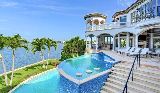 A luxurious Mediterranean-style waterfront mansion with an infinity pool, palm trees, lounge chairs, and a balcony overlooking the water.