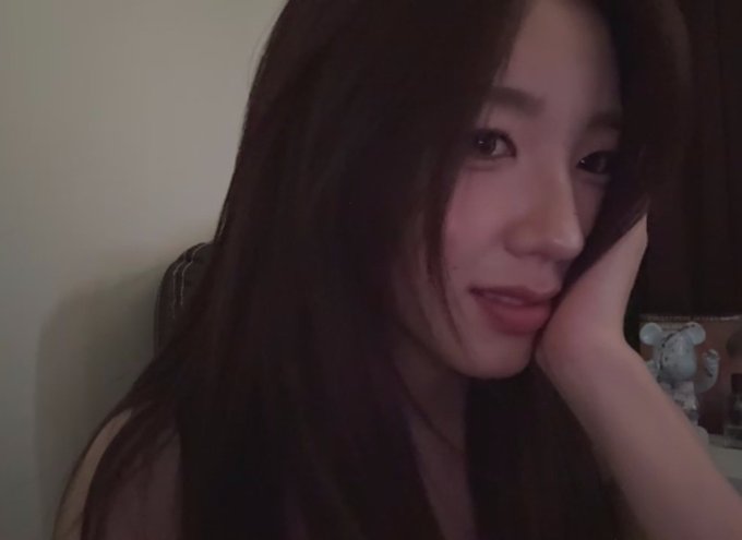 A close-up, dimly lit selfie of a young woman resting her cheek on her hand, smiling softly with a small plush figure visible in the background.