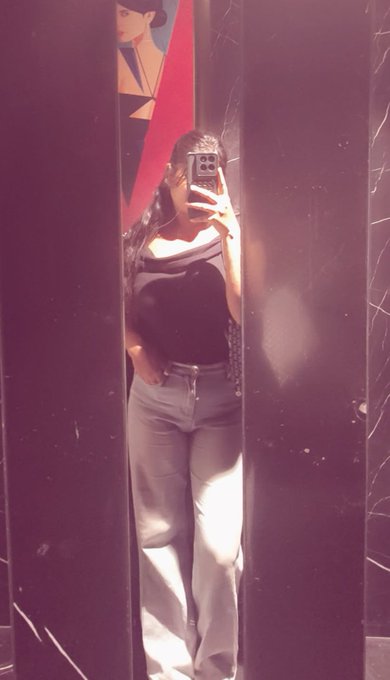 A mirror selfie of a person wearing a black one-shoulder top and light blue jeans, taking a photo with their phone.