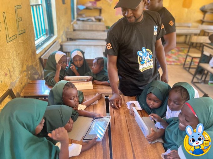 A teacher instructing young students wearing green hijabs who are using tablets in a classroom setting, with a cartoon rabbit logo in the corner.