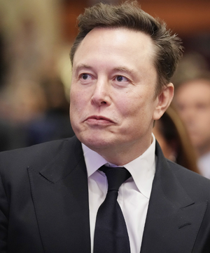 A close-up photo of Elon Musk wearing a black suit and tie with a serious expression. The image accompanies news that his company SpaceX is acquiring nationwide spectrum to expand Starlink into a major US telecom provider, causing carrier stocks like AT&T and Verizon to drop.