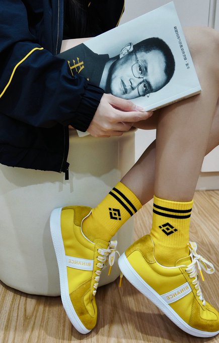 A person sits reading a book featuring a portrait of a bespectacled man on the cover, wearing bright yellow sneakers with matching striped socks and dark jacket.