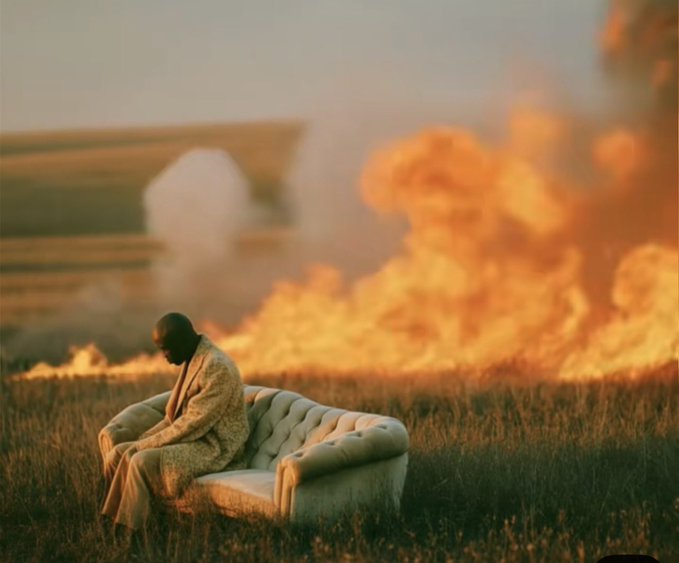 A surreal scene of a man sitting on a white couch in a grassy field with large flames and smoke in the background.