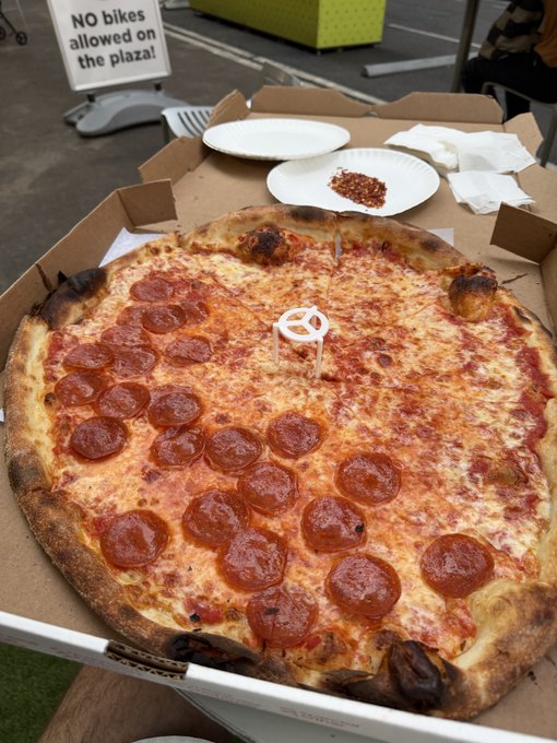A large pepperoni pizza with a half-and-half topping arrangement sits in an open delivery box, accompanied by plates of crushed red pepper flakes and a sign reading "NO bikes allowed on the plaza!" in the background—capturing the triumphant return to enjoying pizza outdoors, true to the "We are so back" sentiment.