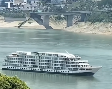 A multi-deck river cruise ship sails on a body of water beneath a bridge, with a town visible on the hillside in the background.