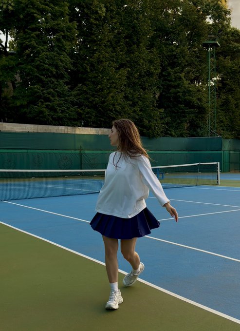 A young woman in a white sweatshirt and navy pleated skirt walks across a blue tennis court, head turned over her shoulder. The athletic setting and sporty outfit contrast with the caption's reflection on gaming, as both sports and video games can spark deep emotional connections that rival any TV show.