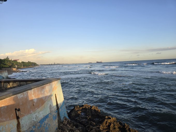 A scenic ocean view at sunset with gentle waves, featuring a ship on the horizon and a weathered concrete structure in the foreground.