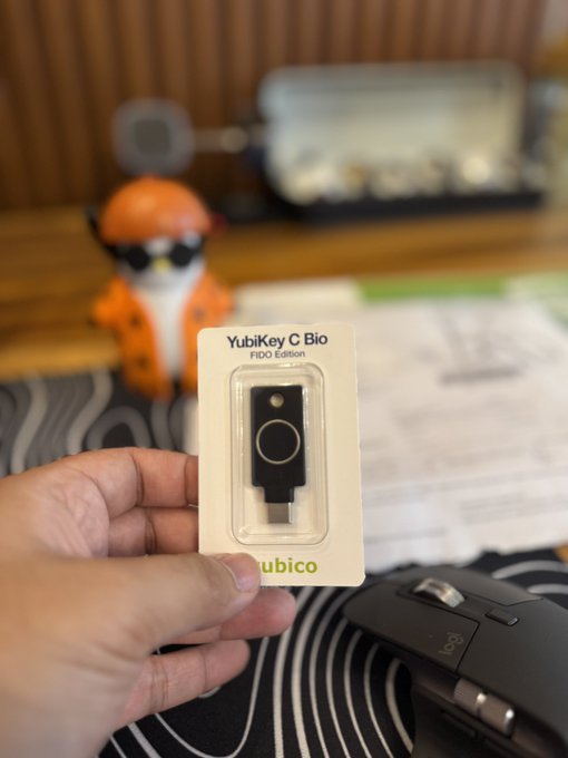 A hand holds a YubiKey Bio FIDO Edition security key in its packaging, with a Logitech mouse and a small orange fox figurine visible in the background.