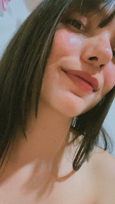 Close-up selfie photo of a young woman with brown hair and bangs, smiling slightly with glossy lips, against a plain light background.
