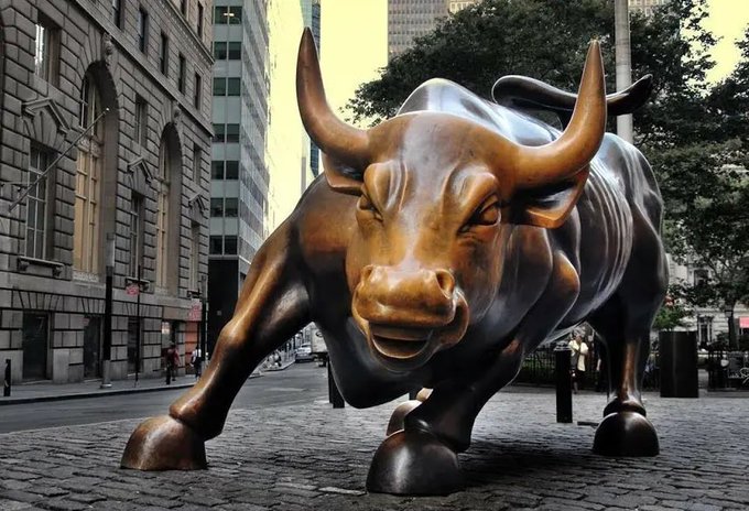 The iconic Charging Bull bronze statue positioned on a cobblestone street in New York's financial district with skyscrapers in the background.