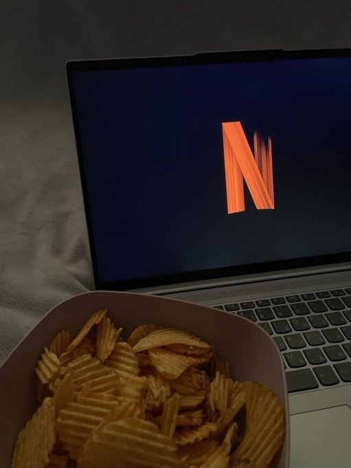 A dimly lit photo of a laptop displaying the orange Netflix 'N' logo, with a bowl of crinkle-cut potato chips in the foreground.