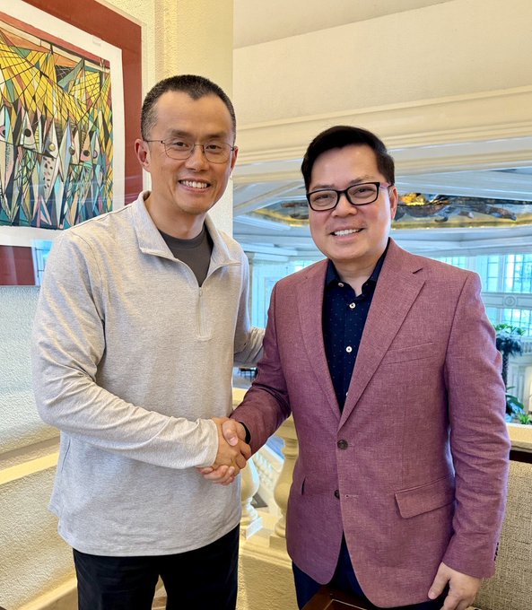 The image shows two men smiling and shaking hands in what appears to be an upscale lobby or hotel interior, with a colorful abstract painting visible in the background. This warmly formal greeting aligns with the post's "See you all tmr, in Manila," suggesting a friendly pre-event meetup, likely ahead of a speaking engagement or business meeting in the Philippines.