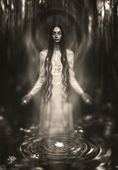 A pale, ethereal woman with long dark hair floats above a pond, surrounded by a glowing aura in a dark, misty forest.