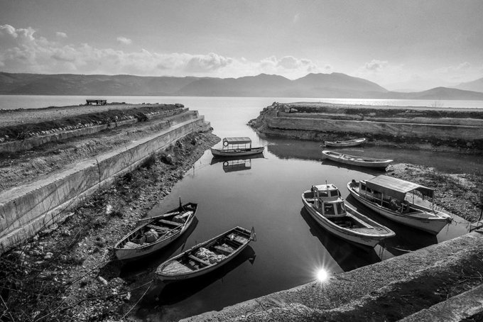 A black and white photograph of a serene harbor with several small boats moored in calm water, framed by stone walls, with mountains visible in the distance and sun reflecting off the water's surface.