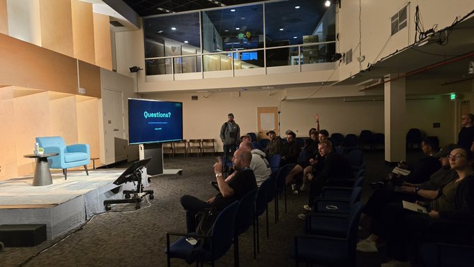 A panel discussion or Q&A session in a modern auditorium, with the screen displaying "Questions?" as an audience member raises their hand.