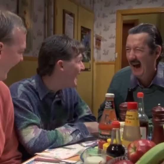 A scene from a sitcom showing three men laughing together at a table with various condiment bottles, fruit, and other items.