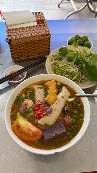 A bowl of Vietnamese noodle soup topped with fried tofu, blood pudding, tomatoes, and chili, served with fresh herbs, bean sprouts, and limes on the side.
