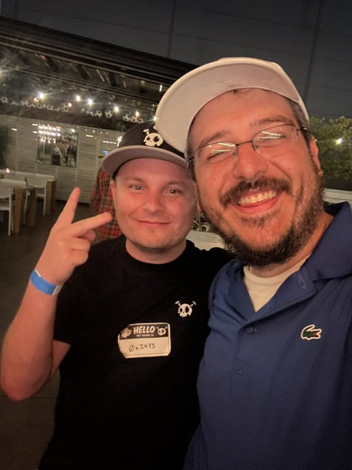 Two men wearing hats (one with a panda logo) pose for a selfie, with one wearing a 'HELLO my name is' sticker and the other in a blue Lacoste polo.