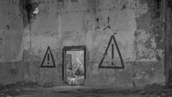 A black and white image shows a weathered, crumbling wall with a broken doorway revealing a glimpse of overgrown ruins beyond; two large warning triangles with exclamation points are painted on either side of the doorway, reinforcing the post's message about the "brutality of indifference" through the symbolic caution signs amid decay.