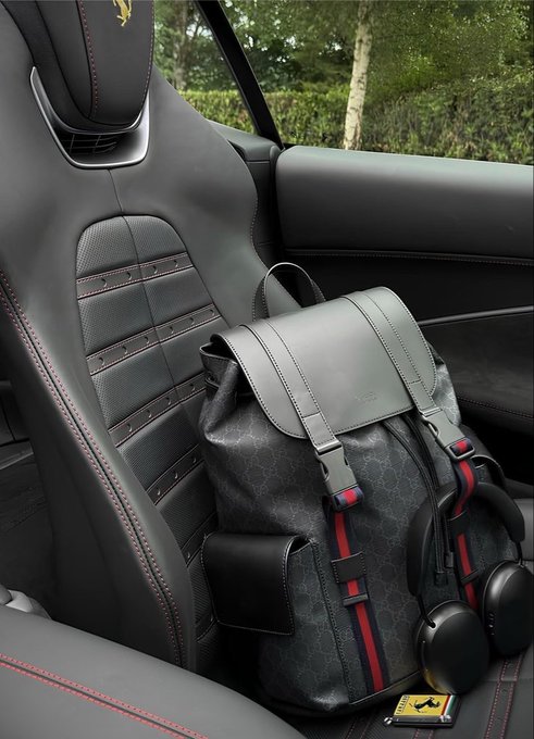 A Ferrari-branded black leather backpack with red, white, and blue striped straps sits on a black leather car seat with red stitching, with Ferrari headphones attached and a small Ferrari prancing horse logo visible.