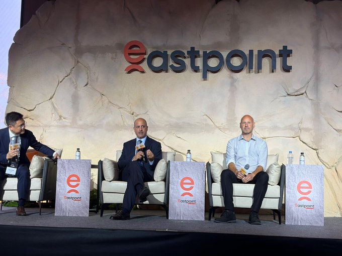 The caption reads: Three men sit on a stage at an Eastpoint event, with one speaking into a microphone while the others listen, in a panel discussion about traditional finance institutions deploying on Ethereum, as mentioned in the accompanying post.