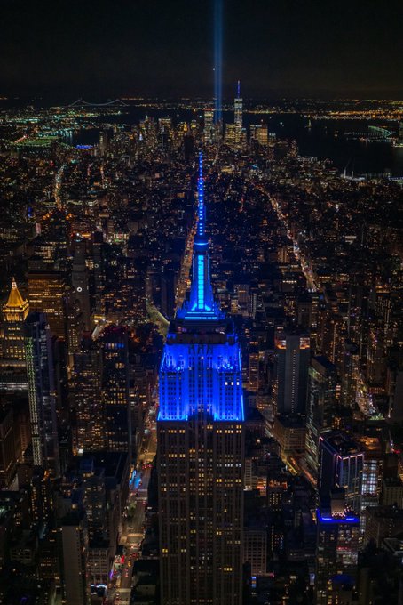 An aerial nighttime view of a city skyline features a central skyscraper illuminated in vibrant blue, with a bright beam of light shooting up from its spire.