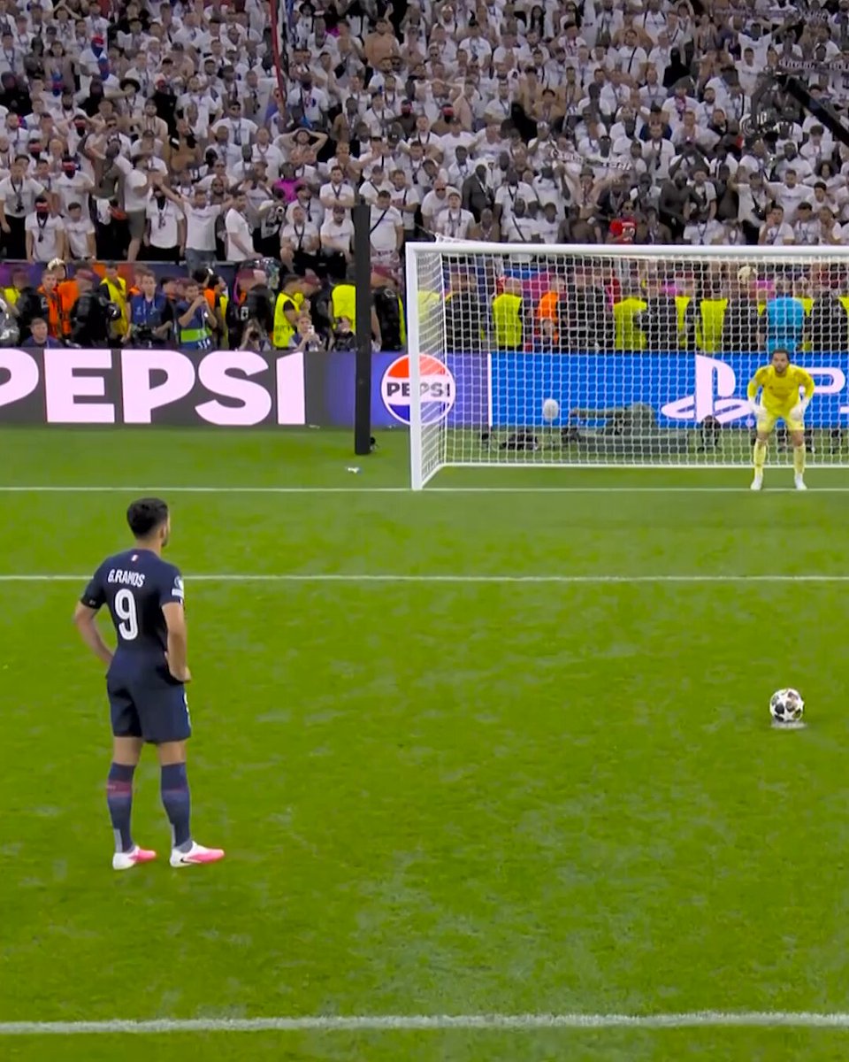 In the image, a soccer player wearing a blue jersey with the number 9 stands near the goalpost, watching as his team attempts to score a goal against another team. The crowd of fans behind him is cheering for their team, creating an atmosphere of excitement and anticipation. The scene captures the intensity of the game and the emotions of both players and spectators alike.