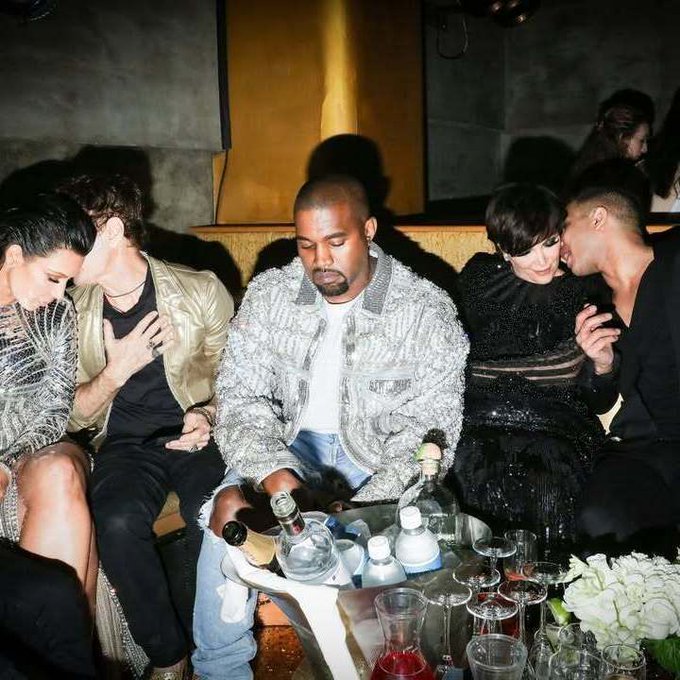 Kanye West sits at a table with bottles and glasses, surrounded by other celebrities including Kris Jenner and Kim Kardashian in a dimly lit setting.