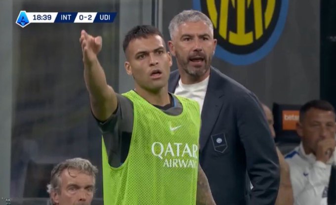 A player in a green "QATAR AIRWAYS" warm-up bib gestures animatedly alongside a suited man during a Serie A match shown on the scoreboard as INT 0-2 UDI at 18:39, illustrating Inter Milan's frustration as assistant manager Aleksandar Kolarov was sent off for protesting a denied penalty.