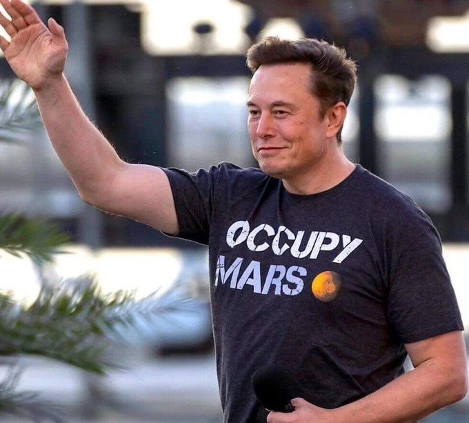 Elon Musk waving while wearing a dark grey t-shirt that reads 'OCCUPY MARS' with a small Mars planet graphic, photographed outdoors in front of a blurred building.