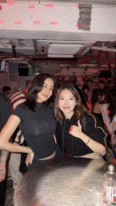 Two stylish women pose together at a trendy, neon-lit venue, with one giving a thumbs-up while wearing a designer jacket featuring "VERSACE JEANS COUTURE" branding—suggesting a luxurious lifestyle that contrasts with the trading-themed caption implying easy market profits. The fashionable scene and high-end accessories visually reinforce the post's sarcastic message about the glamorous facade often presented in trading circles.
