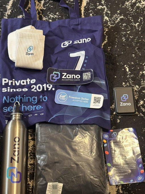 A collection of Zano cryptocurrency merchandise laid out on a dark surface, including a blue tote bag reading 'Zano Private since 2019. Nothing to see here. ZCASH FORKS WIN', a pair of white socks, a Zano-branded sticker and 'Accepted Here' sign, a metal water bottle, a black garment, and a Zano trading card.