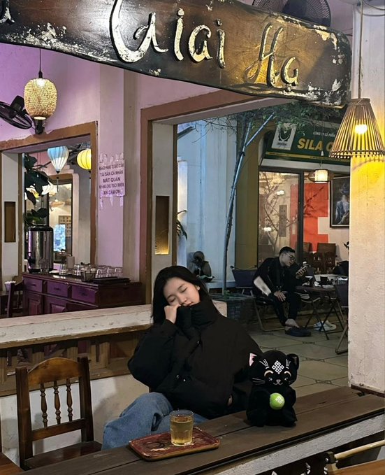 A woman in a black puffer jacket rests her head on her hand at a wooden table in a cafe or bar, with a glass of amber drink and a black lucky cat figurine in front of her.