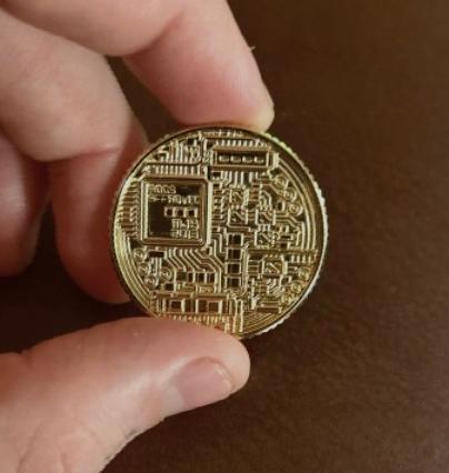 A close-up of a hand holding a physical Bitcoin commemorative coin with visible circuit board engravings and a microchip design on its surface.