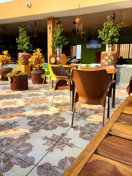 An outdoor café area features brown plastic chairs and green chairs around wooden tables, surrounded by potted plants and decorated with intricately patterned floor tiles.