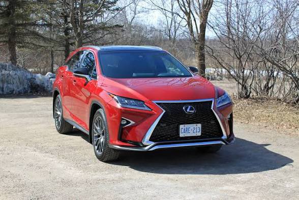 The image features a red Lexus car parked on a dirt road next to some trees. The car appears to be an older model, possibly a Lexus ES-150 or similar make. It has a license plate that reads "CAER-2". The scene is set in a wooded area with a clear blue sky overhead.