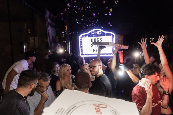 A lively night celebration unfolds under a glowing "PHD DOGS STREET FESTIVAL" marquee, where a crowd raises their phones and confetti rains down as someone hoists a pizza box above the festivities. The scene captures the electric atmosphere of DDNYC 2026, embodying the "Doginal Dogs to infinity" spirit of community and excitement.
