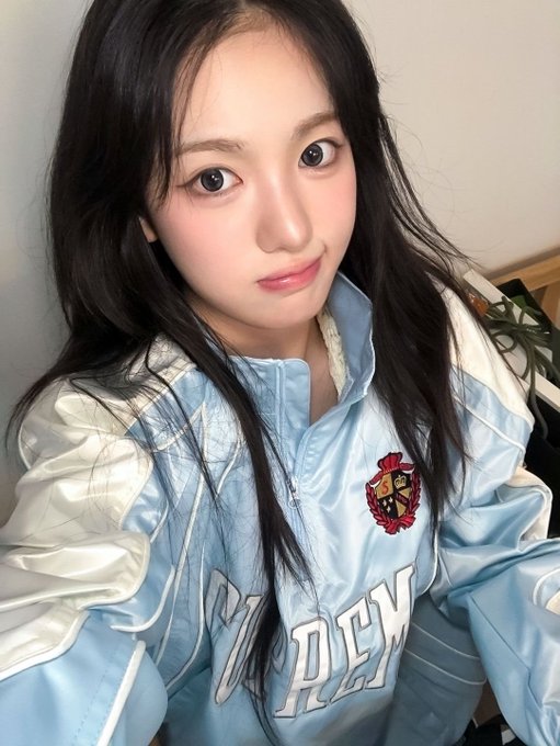 A selfie of a young woman with long dark hair wearing a light blue and white 'Hogwarts' jacket featuring what appears to be a Gryffindor crest.