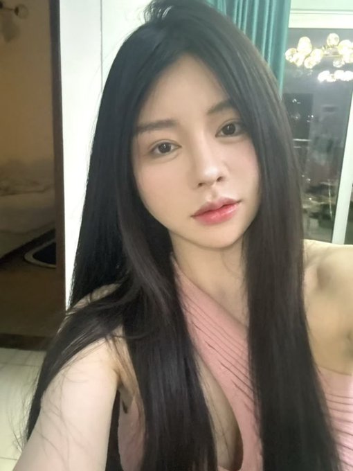 A selfie of a young woman with long dark hair wearing a pink top, taken indoors with teal curtains and a chandelier visible in the background.