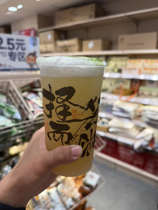 A hand holding a plastic cup of a yellow-green beverage with Chinese calligraphy on it, photographed in what appears to be a Chinese grocery or convenience store.