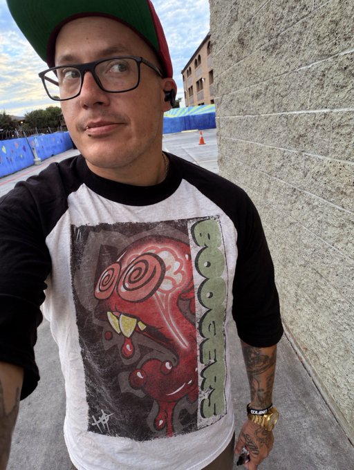 A selfie of a young man wearing glasses, a green and red cap, and a baseball-style shirt with a graphic that reads 'BOOGERMAN', standing next to a concrete wall with blue barriers in the background.
